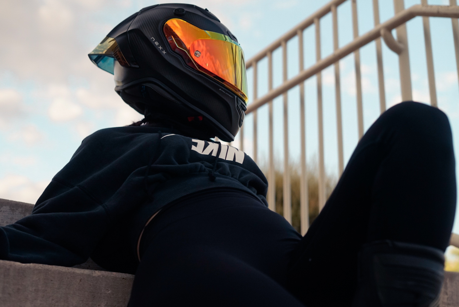 Rider in a black helmet with an orange visor resting against steps under a bright sky