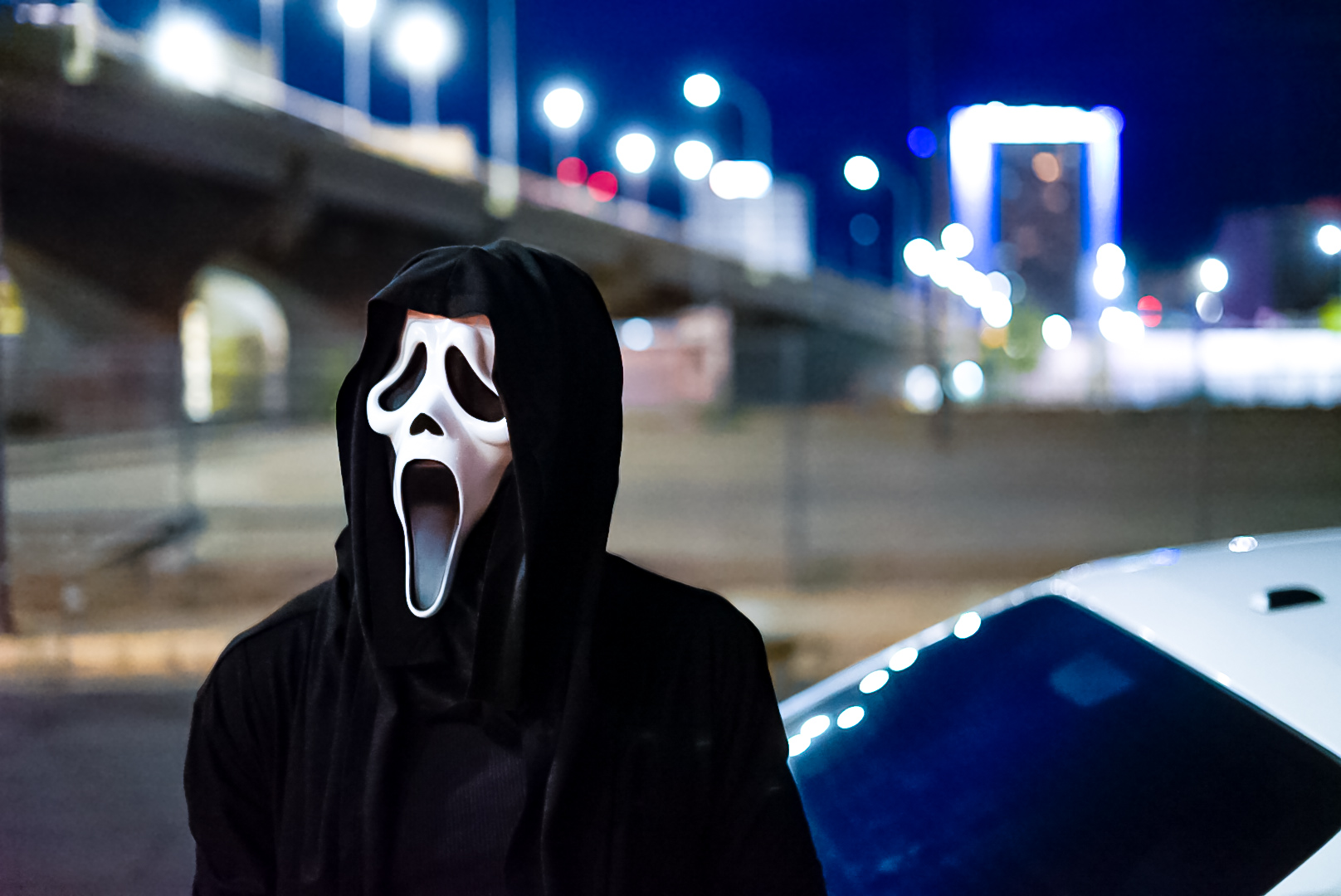 Masked figure in a black hood beside a white car with city lights behind