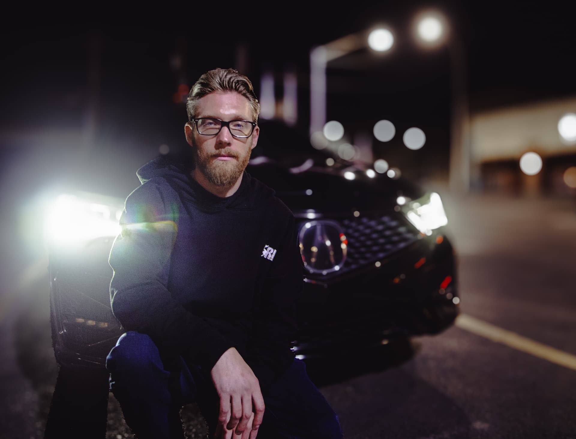 Bearded person in a black hoodie leaning on a car at night