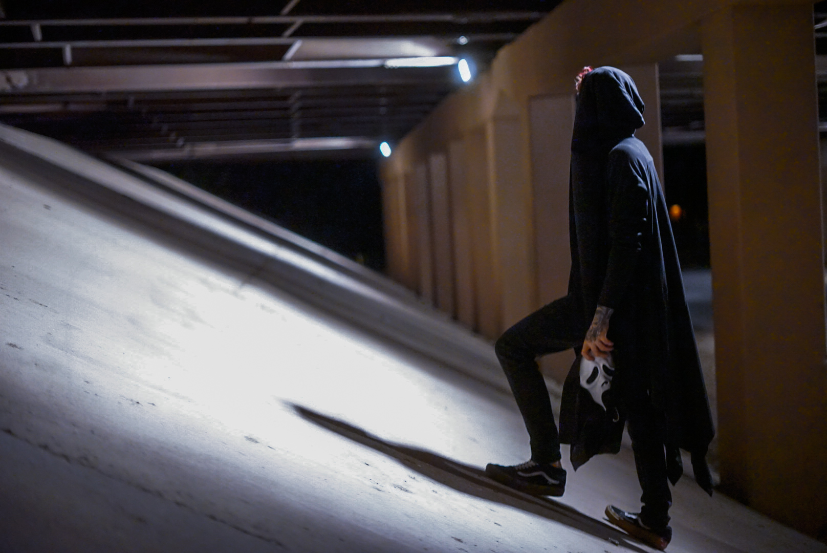 Hooded figure holding a white mask while walking through a lit parking garage