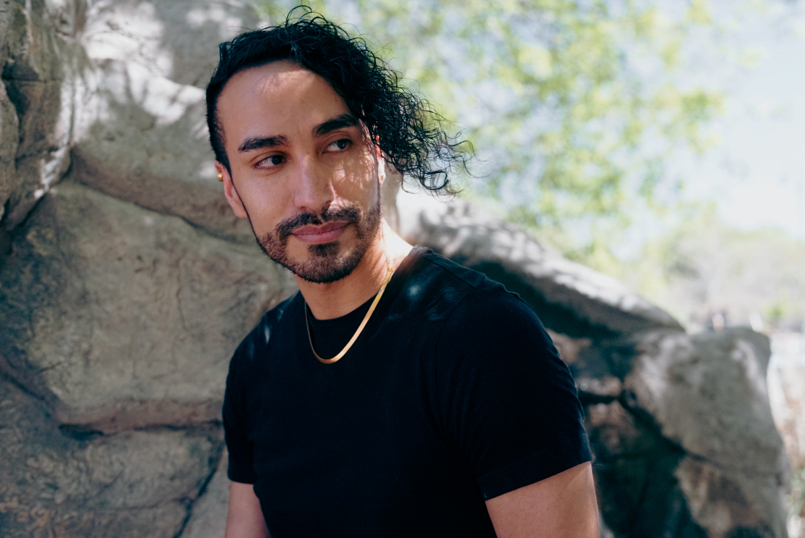 Portrait of a person with curly hair and a gold chain against sunlit rocks