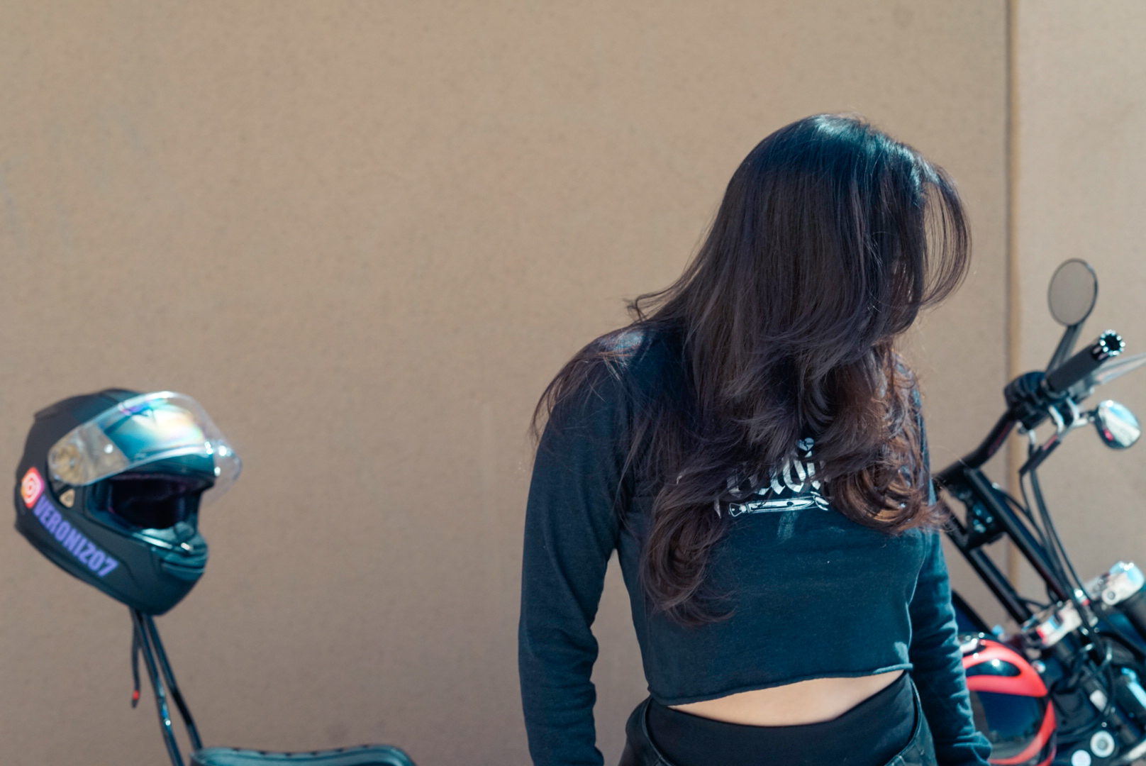 Person with long dark hair beside a motorcycle and helmet against a plain wall