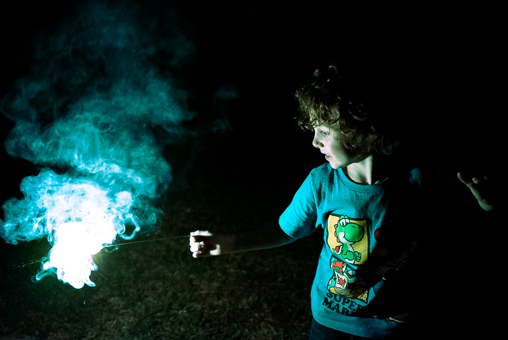 Child holding a sparkler surrounded by glowing teal smoke at night