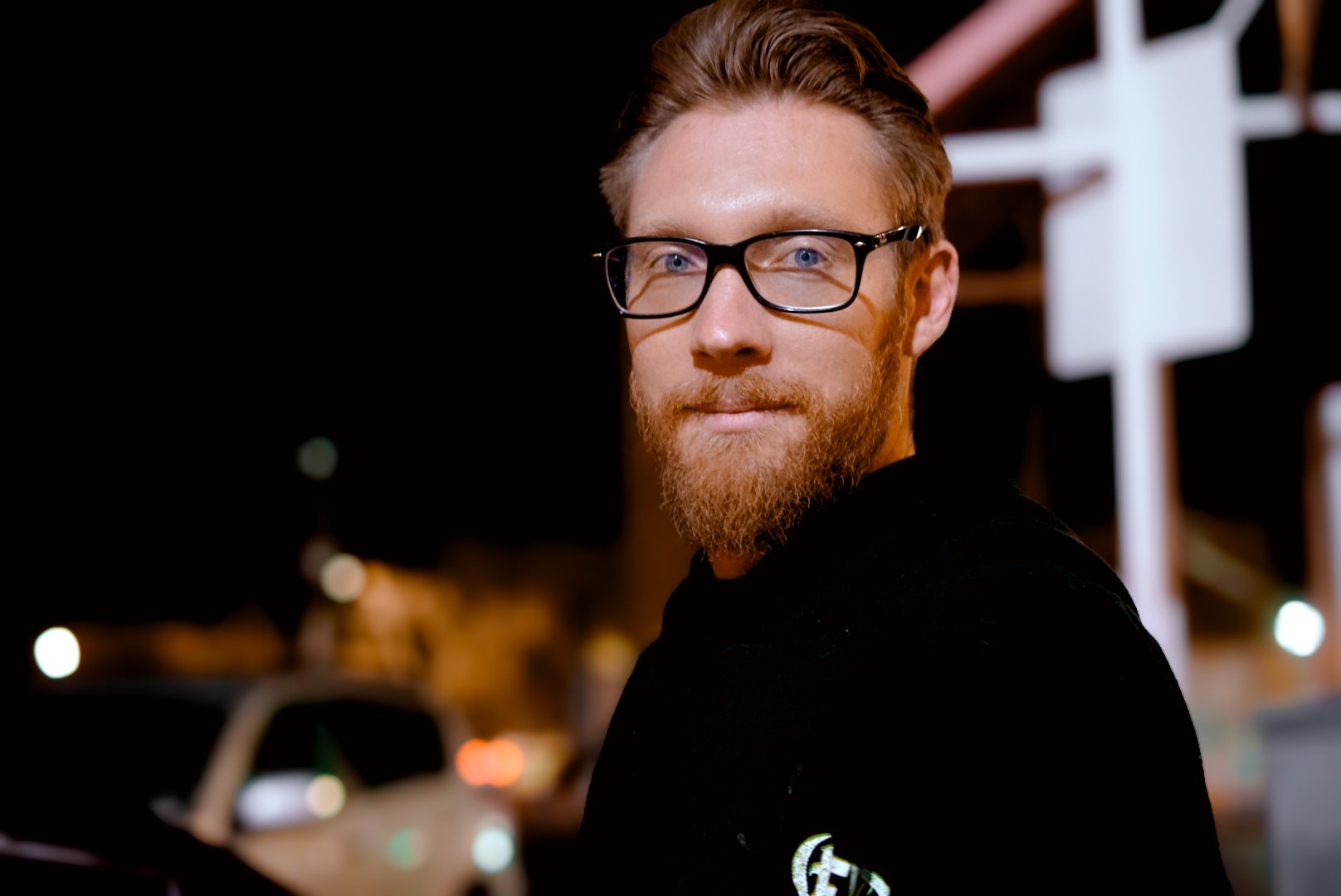 Portrait of a bearded person in glasses at night with city lights behind