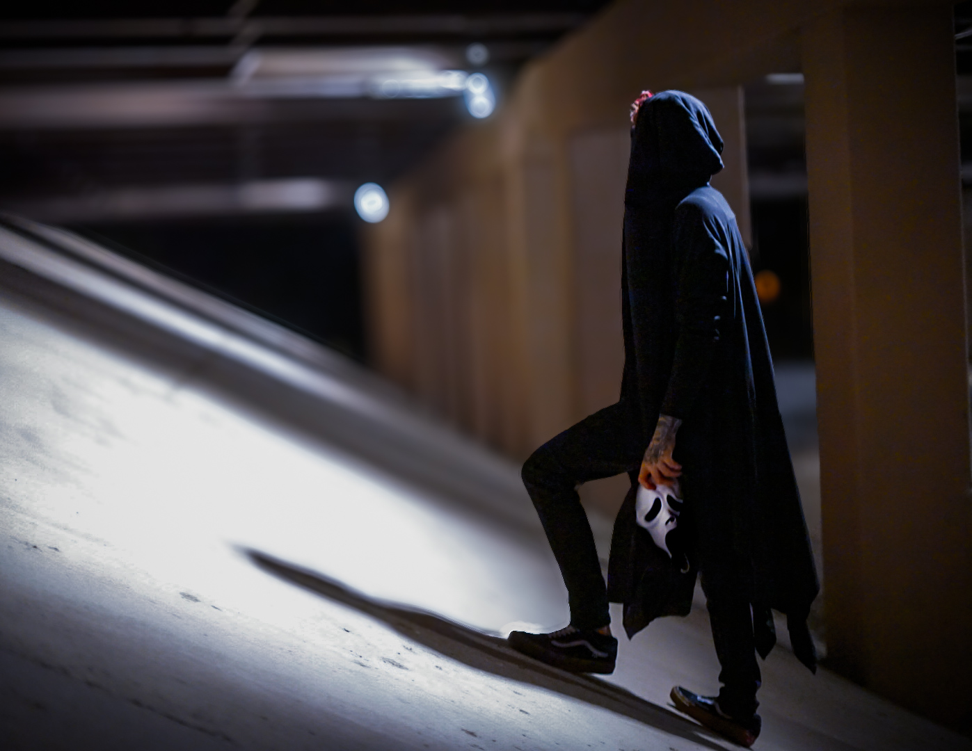 Hooded figure holding a white Ghostface mask while walking through a lit parking garage