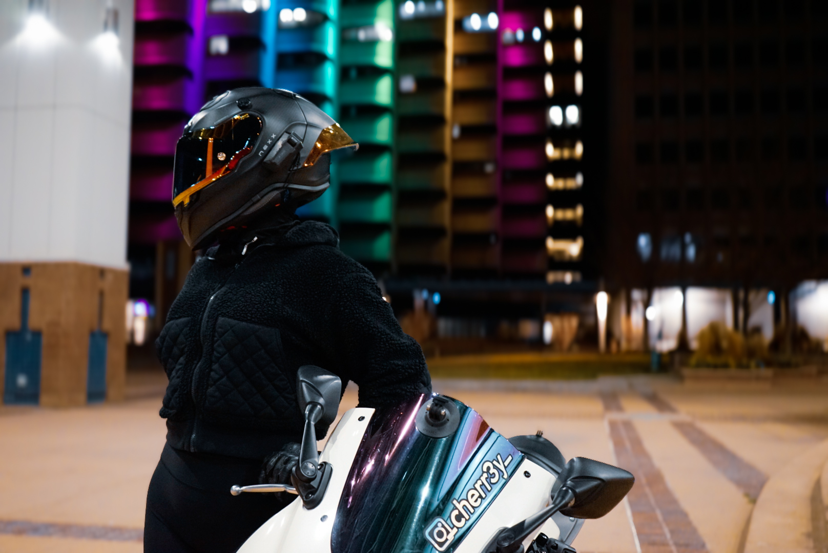 Rider in a black helmet and fleece jacket on a scooter in front of a colorful building at night
